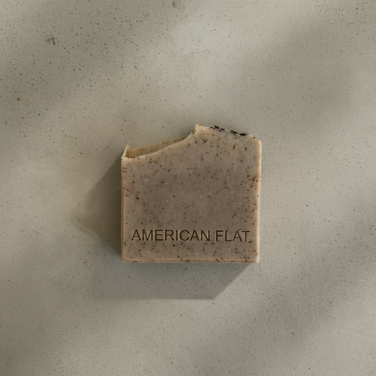 A BROWN BAR OF SOAP SPECKLED WITH GROUND COFFEE STAMPED WITH AMERICAN FLAT ON A CONCRETE BACKDROP