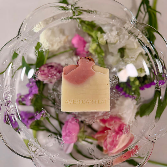 A WHITE AND PINK SWIRLED BAR OF SOAP LAYING ON A GLASS PLATTER WITH DIFFERENT COLORED FLOWERS UNDERNEATH