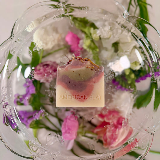 A WHITE, PINK, PURPLE AND GREEN COLORED BAR OF SOAP LAYING ON A GLASS PLATTER WITH DIFFERENT COLORED FLOWERS UNDERNEATH