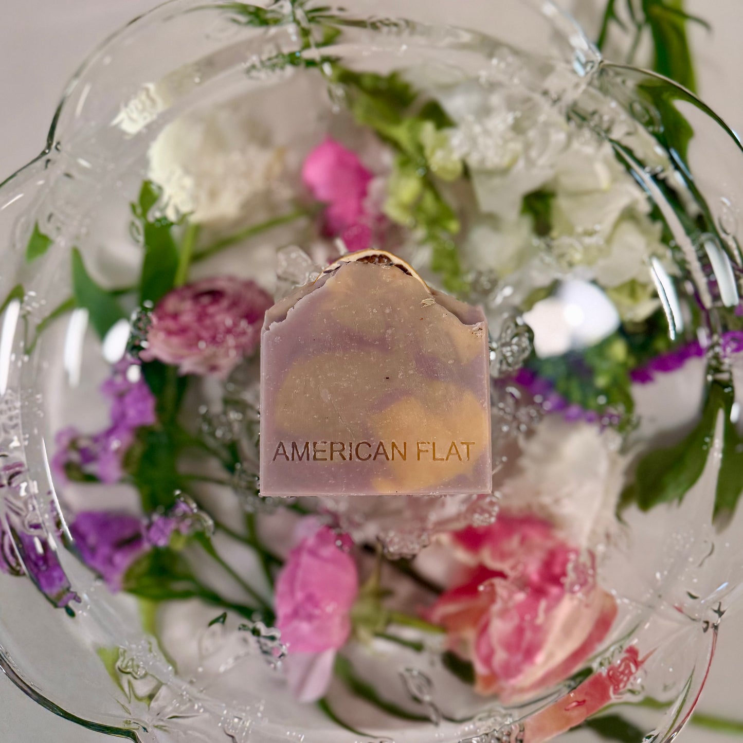 A PURPLE, GREEN AND YELLOW SWIRLED BAR OF SOAP LAYING ON A GLASS PLATTER WITH DIFFERENT COLORED FLOWERS UNDERNEATH