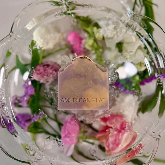 A PURPLE, GREEN AND YELLOW SWIRLED BAR OF SOAP LAYING ON A GLASS PLATTER WITH DIFFERENT COLORED FLOWERS UNDERNEATH