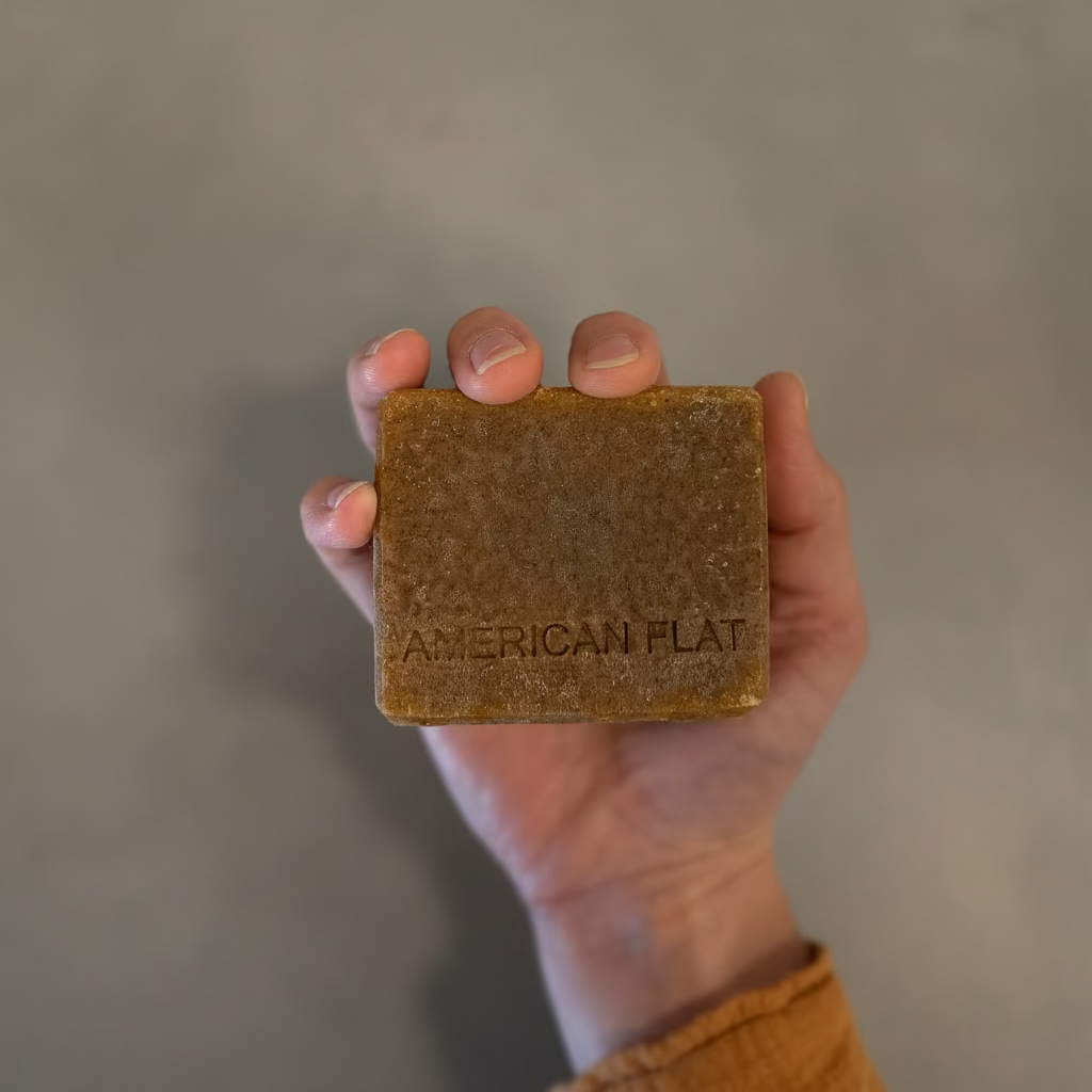 A hand holding a rectangular bar of American Flat soap, which has a textured, earthy brown color, against a blurred gray background.